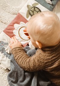 Pastellivärinen puuvillainen aktiviteettimatto, jossa on peili, eläinkuoseja ja erilaisia tekstuureja. Lapsi tarttuu harmaaseen pehmeään leluun samalla kun leikkii.