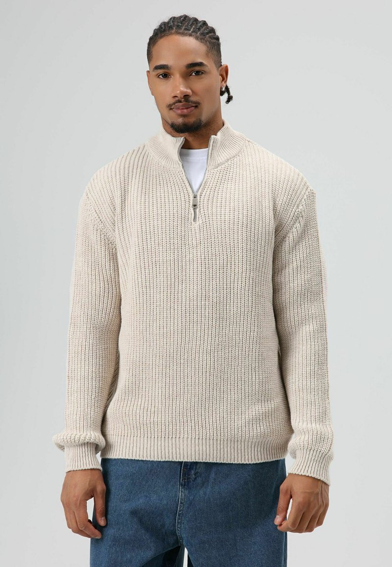 Man met gevlochten haar, gekleed in een crèmegekleurde gebreide trui met halve rits over een wit shirt en blauwe jeans, staand tegen een effen lichte achtergrond.