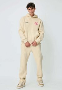 Sweat à capuche en coton beige avec poche avant et broderie de fleur rose, associé à un pantalon beige assorti avec des accents de couture et des baskets blanches.