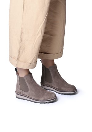 Taupe suede ankle boots with elastic side panels, white stitching, and a rubber sole, paired with beige cropped pants.