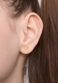 Złota kolczyk w kształcie gwiazdki, umieszczona na tle skóry, prezentująca gładką, błyszczącą powierzchnię oraz minimalistyczny design, blisko ucha.