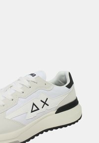 Zapatilla blanca que combina cuero y textil, con un logo geométrico, acentos en negro, suela gruesa y un acabado texturizado en la superficie.