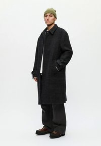 Μακρύ μαύρο παλτό με καρό σχέδιο, φαρδιές πέτρες, ριγμένους ώμους και πλαϊνές σχισμές, σε συνδυασμό με γκρι παντελόνια φαρδιάς γραμμής και καφέ παπούτσια. Πράσινο σκουφάκι στο κεφάλι.