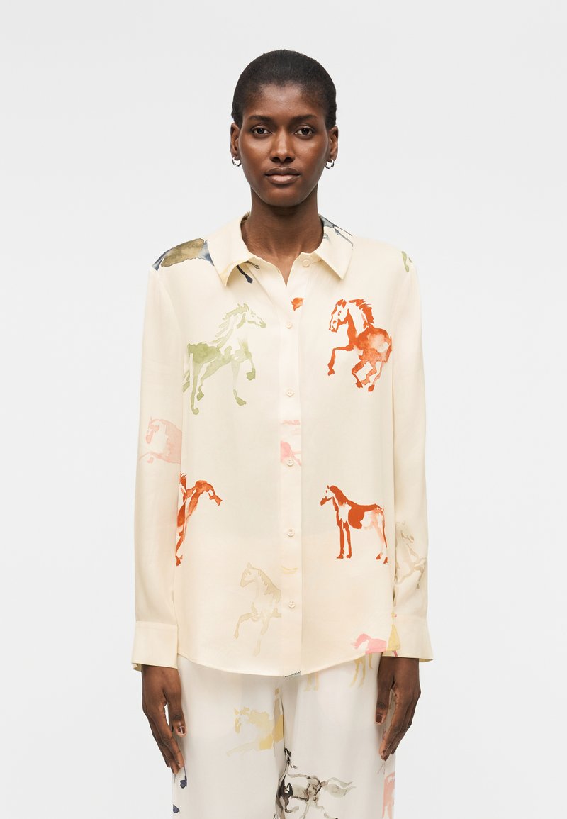 Nainen, joka käyttää kermaan vivahtavaa napillista paitaa ja monivärisiä hevosenkuvioisia housuja, seisoo yksinkertaisen vaalean taustan edessä.