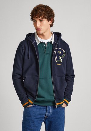 Navy zip-up hoodie with kangaroo pockets, striped orange and green ribbed cuffs, and printed emblem on the chest, layered over a green polo.