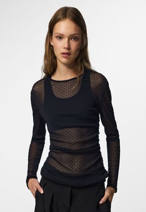 Jeune femme aux longs cheveux portant un top noir transparent à pois à manches longues par-dessus un débardeur noir et un pantalon noir, les mains dans les poches.