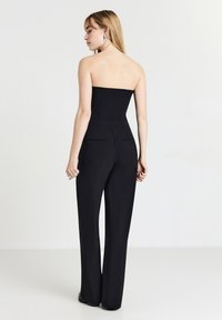 Salopette nera senza maniche con vestibilità su misura, gambe dritte e tasche posteriori. Tessuto dalla trama liscia. Modello che indossa orecchini di argento oversize.