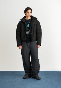 Giacca a vento nera con colletto alto, indossata sopra una felpa scura con grafiche e righe turchesi, abbinata a pantaloni neri larghi.