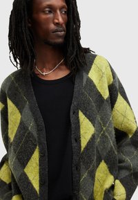Cardigan argyle grigio e giallo in lana strutturata, con chiusura a bottoni e vestibilità ampia, indossato sopra una camicia nera.