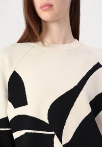 Jeune femme portant un pull beige avec un grand motif floral abstrait noir, vue du cou jusqu'au milieu du torse sur un fond uni.