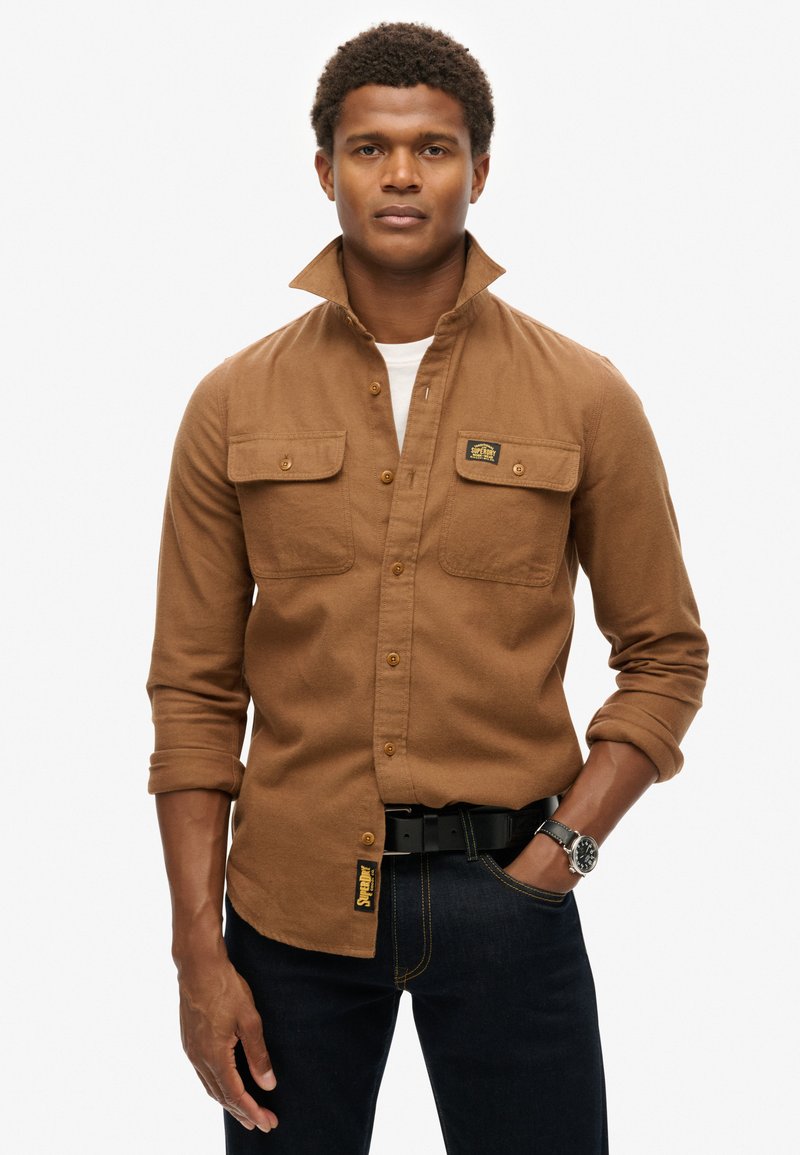 Bruine overhemdd met zachte stof, voorzien van twee borstzakken, opgerolde mouwen en een logo patch. Draag het met een donkere jeans en een zwarte riem.