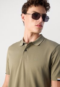 Olíva zöld polo ing texturált felülettel, két gombos nyakkal, fekete gombokkal és egy diszkrét BOSS logóval. Vékony csíkos kiemelések az ujjakon.