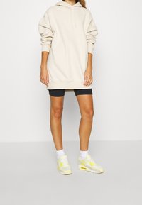 Vestido sudadera beige con capucha y mangas extragrandes, combinado con pantalones cortos de ciclismo negros y zapatillas gris claro con detalles amarillos.