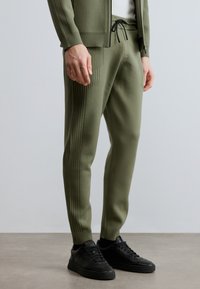 Pantalones de chándal de punto de color verde oliva con detalles acanalados verticales, cintura elástica y cordón negro, combinados con zapatillas negras.