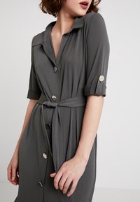 Šedé šaty s krátkým rukávem, límcem, zapínáním na knoflíky a vázáním v pase. Obsahují knoflíky v stříbrném tónu a mají hladkou texturu látky.