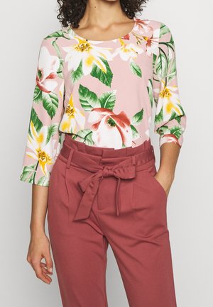 Kobieta ubrana w różowy kwiecisty top z długim rękawem, zestawiony z wysokim stanem mauve spodni z przewiązanym paskiem w talii.