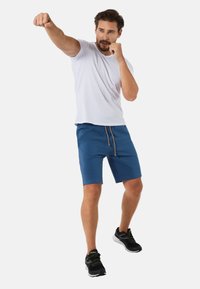 Męska biała koszula krótki rękaw i niebieskie spodenki ze sznurkiem, w pozycji bokserskiej, w czarnych butach sportowych.