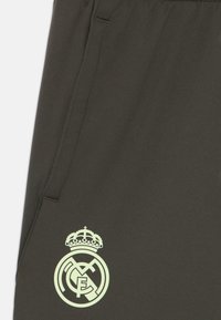 Joggers sportifs vert foncé avec une texture lisse, arborant un logo clair du Real Madrid sur la cuisse gauche et un détail de poche latérale.