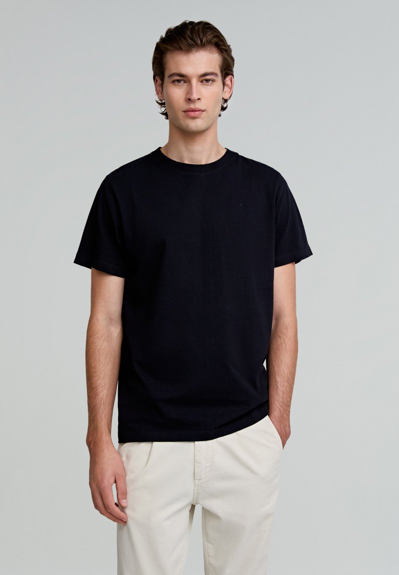 Tricou negru din bumbac cu guler rotund și mâneci scurte, având un detaliu subtil cu logo pe piept, asortat cu pantaloni de culoare deschisă.