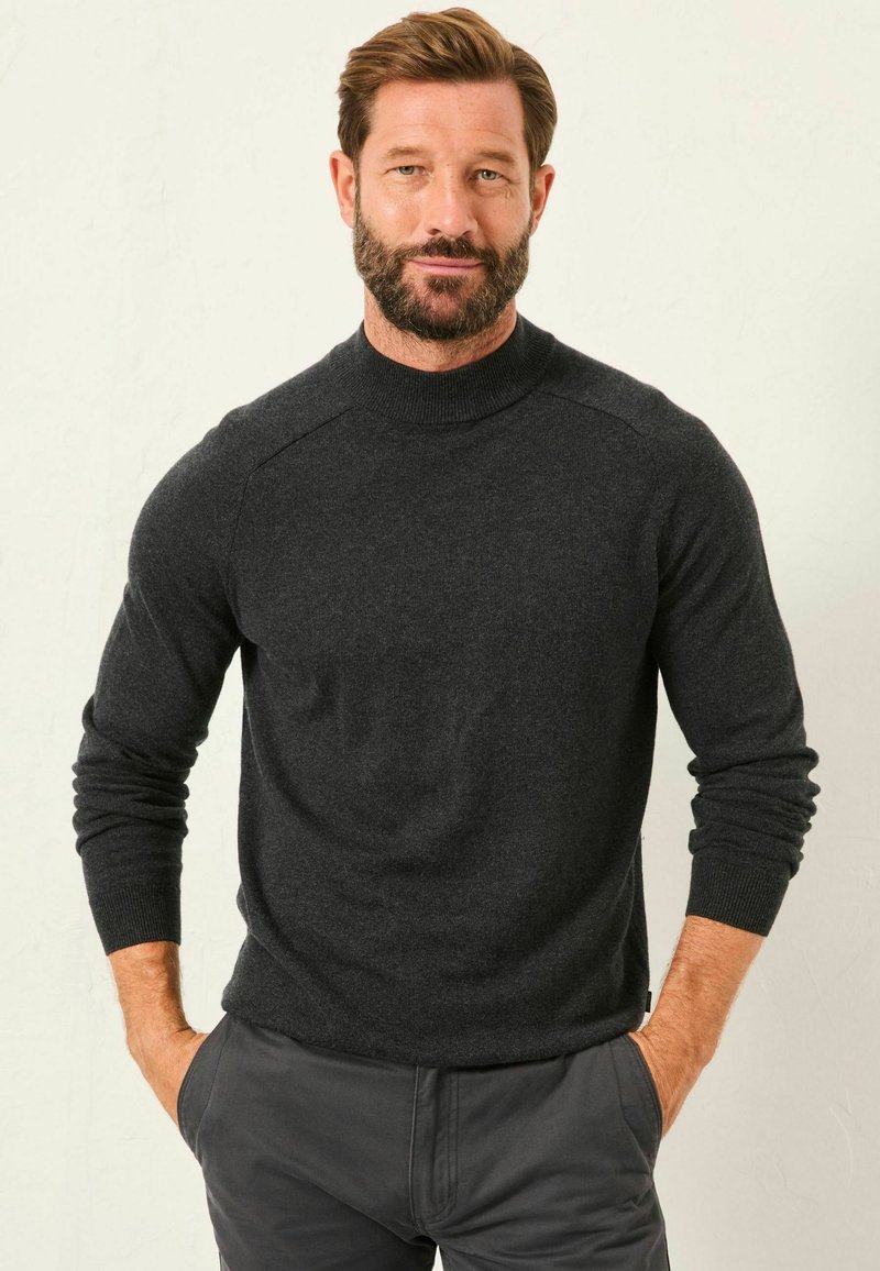 Dunkelgrauer, gestrickter Pullover mit hohem Kragen und gerippten Bündchen. Hat eine lockere Passform und eine glatte Textur. Über grauen Hosen getragen.