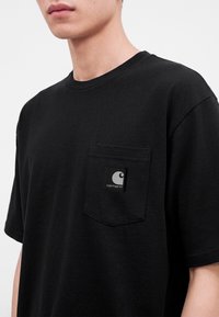 Νεαρό άτομο φοράει μαύρο κοντομάνικο t-shirt Carhartt με τσέπη στο στήθος και λογότυπο στη αριστερή πλευρά.