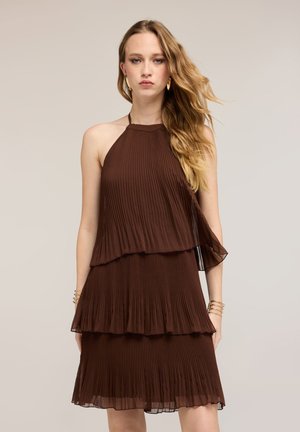 Junge Frau mit langen, welligen Haaren, die ein ärmelloses, gestuftes, plissiertes braunes Kleid trägt, dazu goldene Creolen und Armbänder.