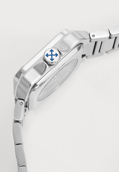Vue latérale d'une montre-bracelet en métal montrant une couronne avec un dessus blanc et un symbole de flèche bleue à quatre directions, attachée à un bracelet métallique à maillons.