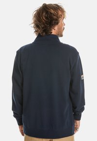 Navy pullover jacket with a high collar, long sleeves, and ribbed cuffs. Features a logo patch on the right sleeve. Soft fabric texture.