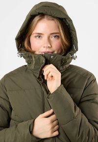 Olijfgroen pufferjack met capuchon, voorzien van een ritsluiting en drukknoopjes. De stof is zacht met een gewatteerd ontwerp en verstelbare trekkingkoordjes.