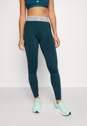 Blue-green leggings with a thick, light gray waistband featuring the Adidas logo. Smooth texture, full-length design, paired with light blue shoes.