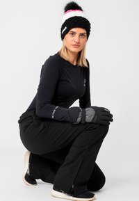 Sort langærmet skjorte, sorte skibukser, grå handsker med en rem, sort og hvid strikhat med pink accenter, i en hukket stilling.