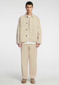 Beżowa bawełniana kurtka z kołnierzykiem, zapinana na guziki z przodu, z kieszenią na piersi, zestawiona z pasującymi beżowymi, luźnymi spodniami i butami.