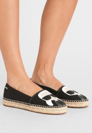 Des espadrilles noires avec un dessus en toile, des éléments de design blancs, un bout rond, une semelle en corde de jute, et un style sans talon à enfiler.