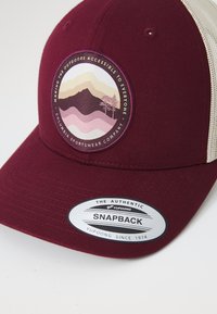 Burgundi snapback sapka, texturált karimával, kör alakú folttal, amelyen pasztell színekben hegyek és fák láthatók, valamint egy ezüst hitelesítő matricával.