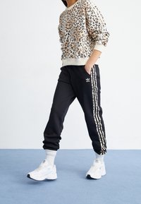 Persona che indossa una felpa con stampa leopardata e pantaloni da jogging neri Adidas con strisce leopardate abbinate, in piedi su un pavimento blu.
