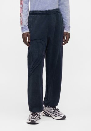 Personne portant un pantalon noir ample avec un motif texturé sur la jambe gauche et des baskets argentées et noires.