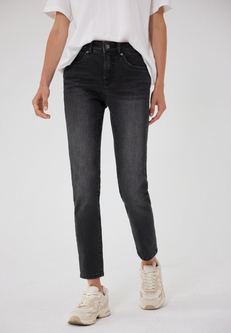 Personne portant un jean slim noir, un t-shirt blanc et des baskets beiges, debout devant un fond clair uni.