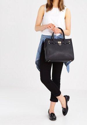 Femme debout tenant un grand sac à main noir, portant un haut blanc sans manches, un pantalon noir retroussé, des mocassins noirs et une veste en denim nouée autour de la taille.