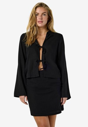 Ženska z dolgimi blond lasmi, oblečena v črno bluzo z dolgimi rokavi in sprednjimi zavezovanji ter ujemajočo se črno mini krilo, stoji pred belim ozadjem.