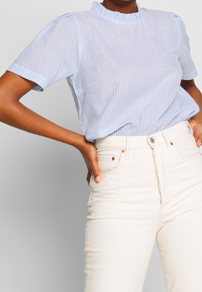 Ljusblå kortärmad blus med vita ränder, volangkrage och mjukt tyg; tillsammans med högmidjade beiga jeans med kopparknappar.