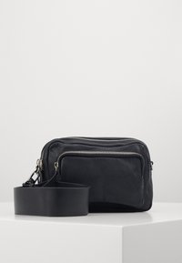 Sorth læder crossbody taske med to lynlåslommer, en aftagelig rem og en struktureret overflade. Kompakt rektangulær form.