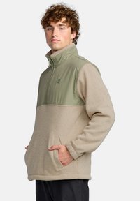 Polarowy sweter z zieloną górną częścią i beżową dolną częścią. Posiada wysoki kołnierz, zamek błyskawiczny oraz boczne kieszenie. Miękka tekstura.