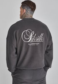 Sudadera gris oscuro de corte holgado, con un gran logo blanco y texto en la parte trasera. Tejido suave, puños acanalados. Diseño casual.