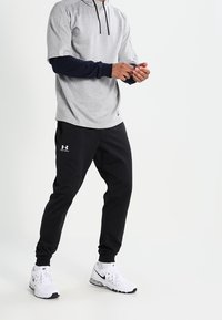 Šedé tričko s krátkým rukávem a kapucí, černé rukávy, doplněné o černé sportovní kalhoty s logem Under Armour a bílé tenisky.