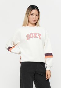 Fehér pulóver "ROXY" logóval korallos színben. Az ujjakon színes csíkos részletek vannak narancssárga és sötétlila színben. Puha, laza dizájn.