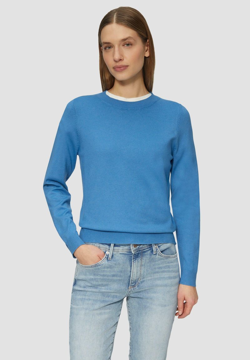 Blauer Baumwollpullover mit langen Ärmeln und rundem Halsausschnitt, mit glatter Textur und tailliertem Saum. Über hellblauen Jeans getragen.