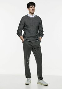 Grå texturerad tröja över en vit skjorta med krage, tillsammans med grå byxor och vita sportskor, stående med händerna i fickorna.