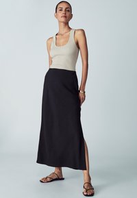 Beige žebrovaný top bez rukávů a černá maxi sukně se bočním rozparkem, nošené s plochými černými sandály, na světlém pozadí. Jednoduchý a moderní design.