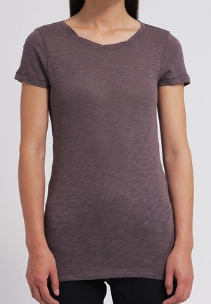 Vrouw die een nauwsluitend donker mauve T-shirt met korte mouwen en zwarte broek draagt, staande tegen een effen lichtgrijze achtergrond.