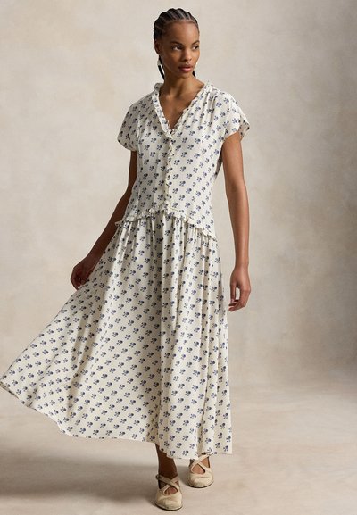 Nainen, joka käyttää kermanväristä kukallista lyhythihaista mekkoa, jossa on röyhelökaulus ja helma, seisoo pitäen toista kättään kohottamassa liehuvaa hametta.